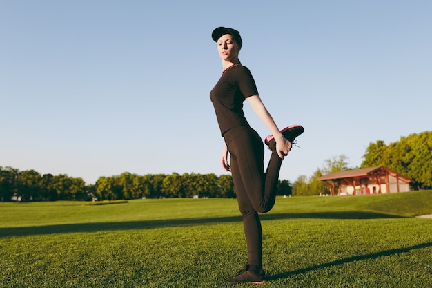 Young athletic girl in black uniform, cap doing sport exercises, warm-up, stretching before running on green lawn in golf course park outdoors on sunny summer day. Fitness, healthy lifestyle concept.