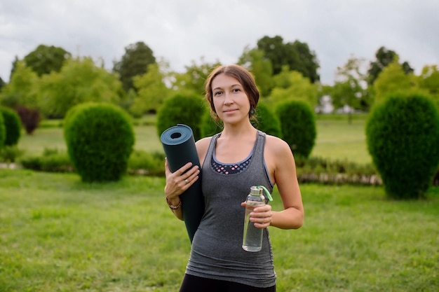 A young athletic girl in a gray tracksuit for fitness is going to do yoga in a green park.