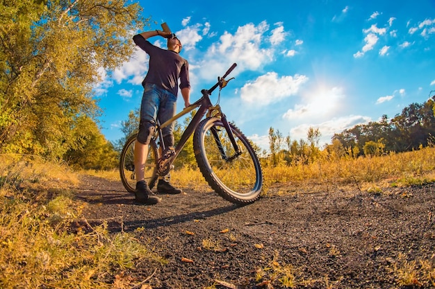 Il giovane uomo atletico in una maglietta nera, pantaloncini di jeans blu e ginocchiere su una bici sportiva beve acqua da uno shaker in un autunno colorato con un cielo luminoso.