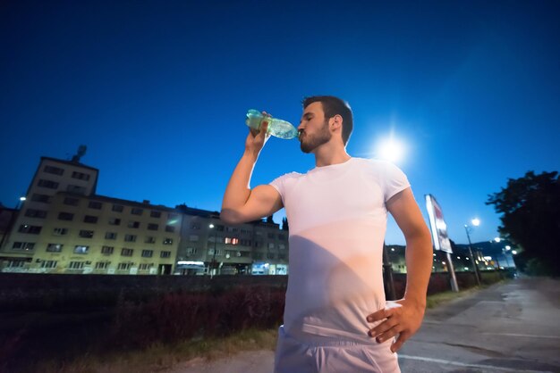 young athletic man drinking water after a night running session in the city