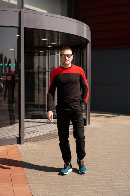 A young athletic man in sunglasses in a tracksuit walks out of the revolving doors of a hotel or shopping mall