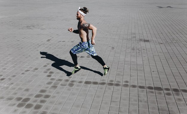 검은색 레깅스와 파란색 반바지를 입은 머리에 문신과 머리띠를 한 벌거벗은 몸통을 가진 젊은 운동선수는 따뜻하고 화창한 날 포장용 석판을 달리고 있습니다.