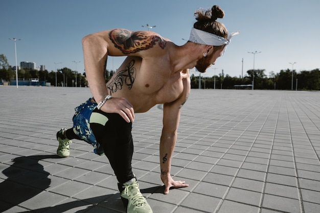 Young athletic man with a naked torso with headband dressed in the black leggings and blue shorts is doing stretching on paving slabs on a warm sunny day .