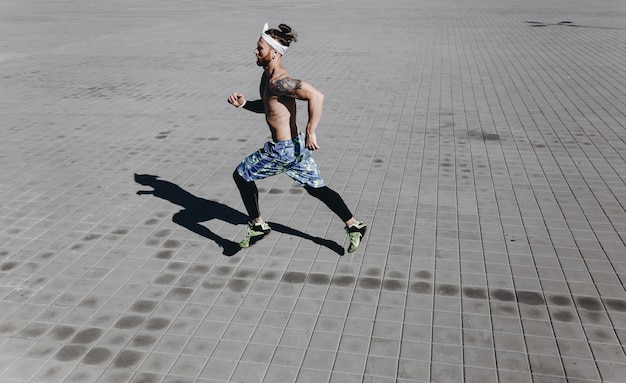 Young athletic man with a naked torso with tattoos and headband on his head dressed in the black leggings and blue shorts runs on paving slabs on a warm sunny day .