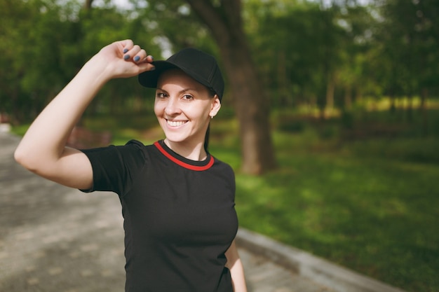 Giovane atletica sorridente bella donna bruna in uniforme nera e berretto in piedi, guardando la telecamera tenendo la mano vicino al cappuccio sull'allenamento sul percorso nel parco cittadino all'aperto