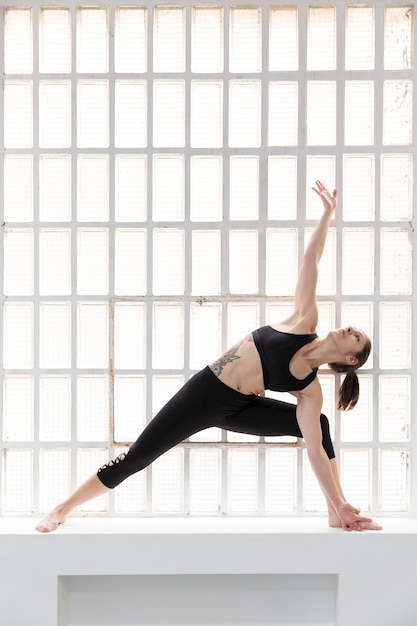 Giovane donna atletica in abiti sportivi neri a praticare yoga accanto a una grande finestra nel suo studio. spazio per il testo.