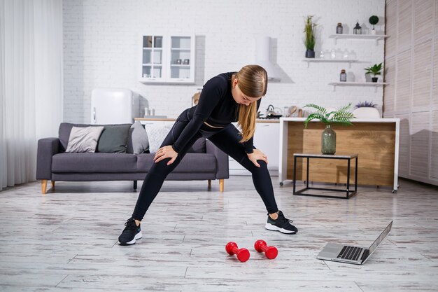 Young athletic woman in fitness clothes black top and leggings in an apartment using online workout from a fitness site on a laptop and doing sports at home. Fitness at home during quarantine