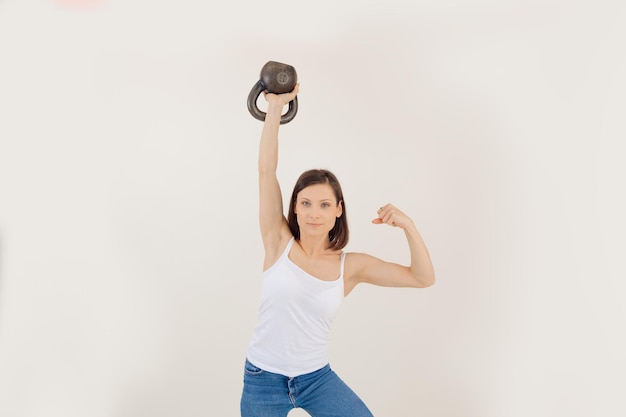Young athletic woman lifting up kettlebell and straining muscles white background keeping fit by str