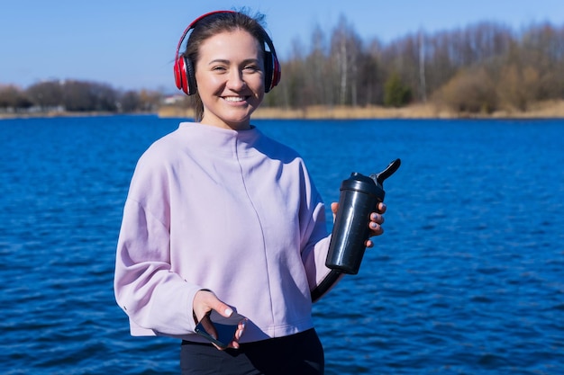 A young athletic woman listens to music and drinks water during breaks from training on the embankment