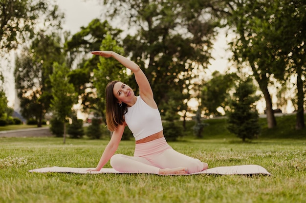 Young athletic woman practice yoga on gymnastic mat in green park on grass Do do side bends and stretch muscles in sportswear in summer morning Concept of healthy lifestyle and harmony with body