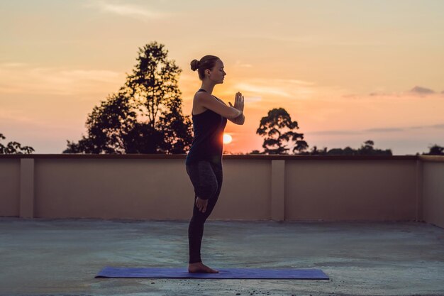 Young athletic woman practicing yoga on the roof at sunset, silhouette.