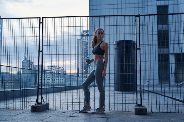 Young athletic woman in sports clothes standing with water bottle in hands on urban city location