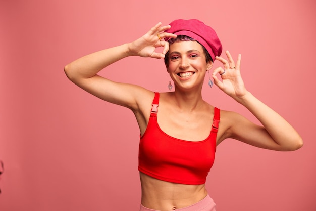 Young athletic woman with a short haircut and purple hair in a red top and a pink hat with an athletic figure smiles and grimaces looking at the camera on a pink background