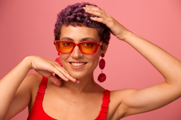 Young athletic woman with a short haircut and purple hair in a red top and pink yoga leggings in sunglasses with an athletic figure smiles and dances on a pink background