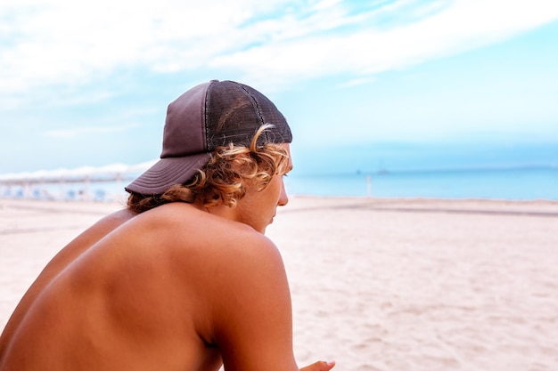 Young attceractive guy surfer sits with his back to the camera on the terrace on the beach looking out into the distance to the see