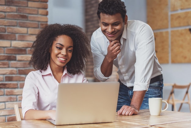 Young attractive AfroAmerican business couple using laptop talking and smiling while working in cafe