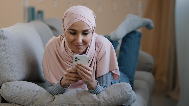 Young attractive arabian muslim girl islamic woman lying on home sofa happy lebanese lady wear hijab