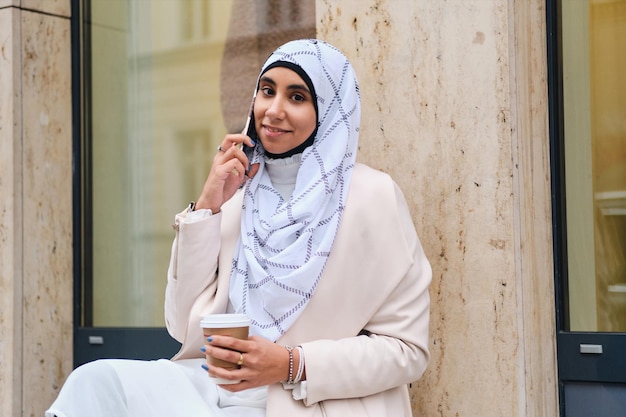 Young attractive Arabic woman in hijab with coffee to go talking on smartphone on city street