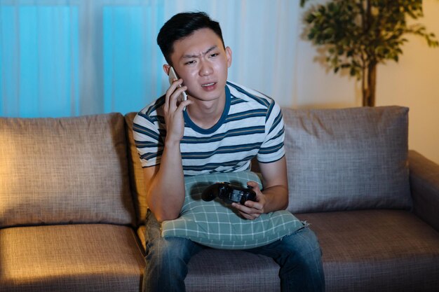 Young attractive asian chinese guy holding joystick and unhappy talking on mobile phone. Serious upset man losing video game in late night evening. confused male discussing with friend on cellphone