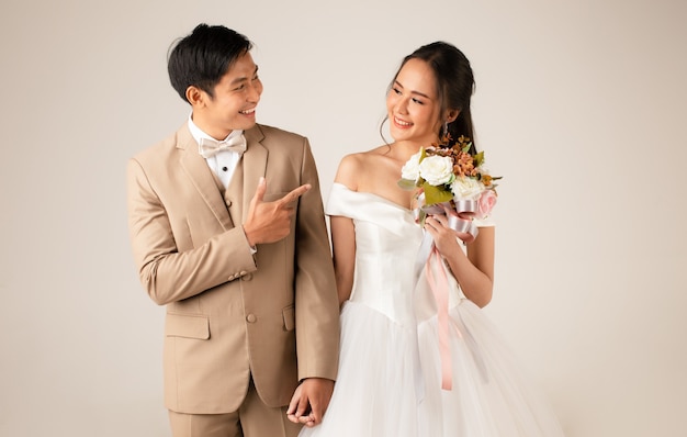 Young attractive Asian couple, man wearing beige suit, woman wearing white wedding gown standing together holding hands. Concept for pre wedding photography.