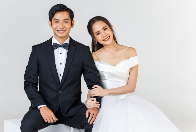 Young attractive Asian couple, soon to be bride and groom, woman wearing white wedding gown. Man wearing black tuxedo, sitting down together. Concept for pre wedding photography.