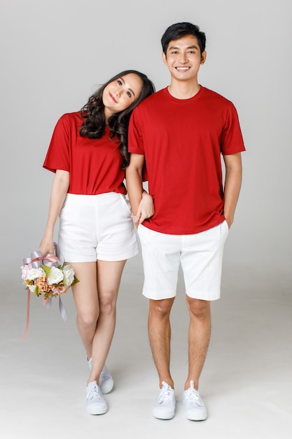 Young attractive Asian couple wearing red t shirt and white shorts standing together, woman holding bouquet of flower against white background. Concept for pre wedding photography. Isolated.