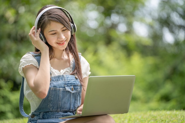 젊은 매력적인 아시아 여성 직원은 이어폰을 끼고 공원에서 온라인으로 디지털 온라인 코스 개발 연구를 듣습니다.