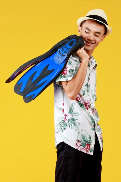 Young attractive Asian man wearing hat and white Hawaiian shirt with red and green flower patterns holding blue flippers with going for beach vacation against yellow background.
