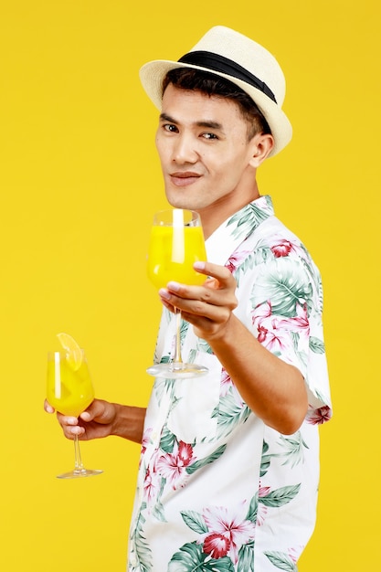 Young attractive Asian man in white Hawaiian shirt holding two glasses of orange juice against yellow background. Concept for beach vacation holiday.