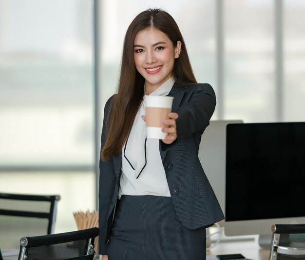 Young attractive Asian woman in black business holding a cup of coffee smiling in modern looking office with blurry windows background. Concept for modern office lifestyle.