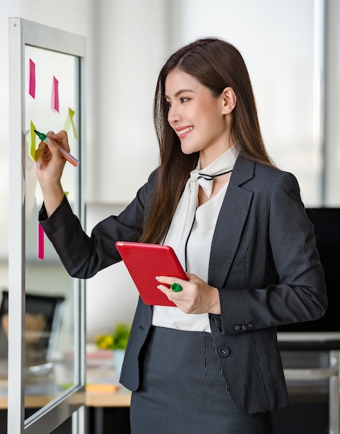 Young attractive Asian woman in black business writing on post it on glass panel one hand holding tablet in modern looking office with blurry windows background. Concept for modern office lifestyle.