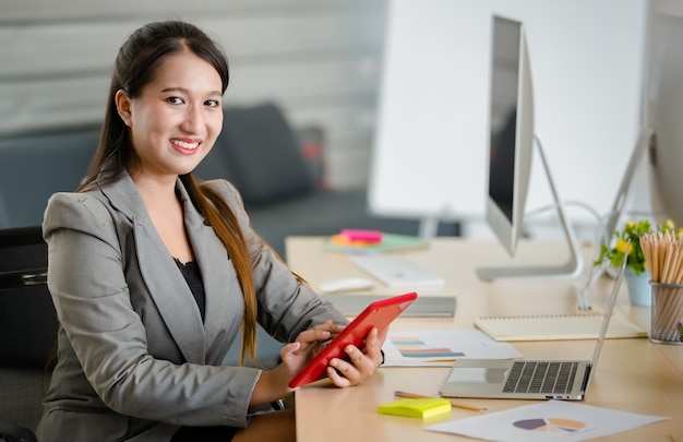 Young attractive Asian woman in grey business suit working on red tablet in modern looking office with blurry windows background. Concept for modern office lifestyle.