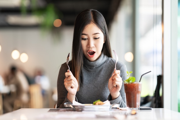 Young attractive asian woman holding fork and spoon feeling hungry