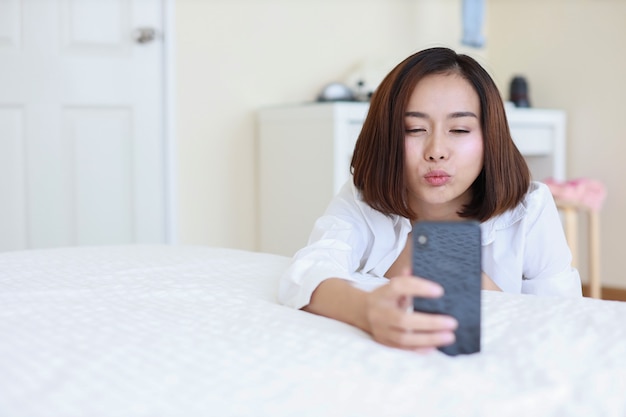 Young attractive asian woman wearing white shirt using VDO call from cell phone and sending a kiss to someone while sitting in  bedroom