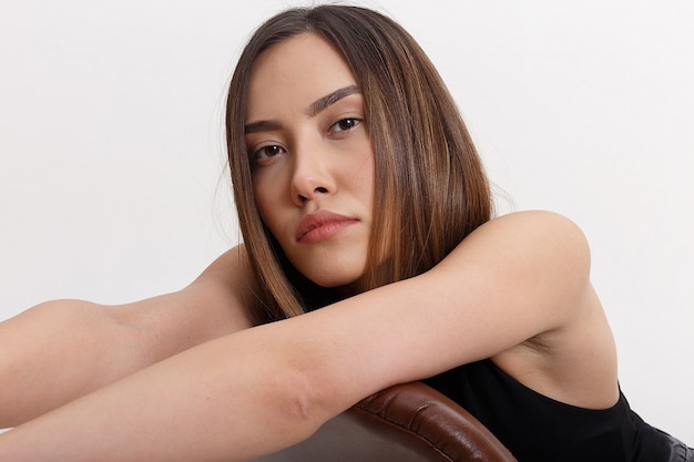 Foto giovane donna asiatica attraente con lunghi capelli castani, isolato sul muro bianco. femmina graziosa magra appoggiata sullo schienale della sedia sul ciclorama. test di modello di bella signora, primo piano