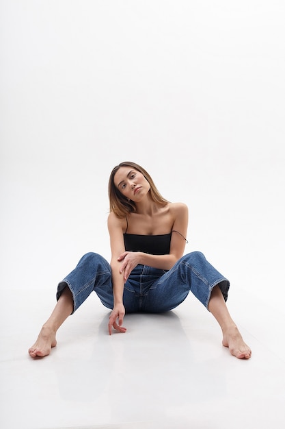 Foto giovane donna asiatica attraente con capelli lunghi in top nero, blue jeans isolato sul muro bianco. magra bella femmina seduta sul cyclorama a piedi nudi. prove modello di bella signora