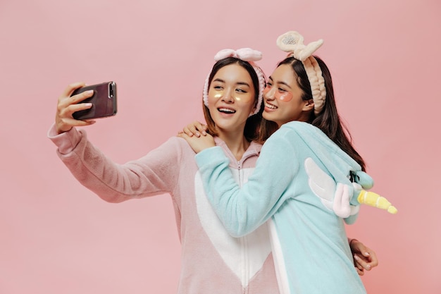 Young attractive Asian women in pajamas take selfie on pink background Happy brunette girls in headbands hug on isolated