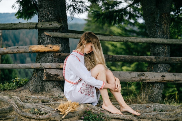 Young attractive barefoot blonde girl in white dress with ornament sitting near wooden fence outdoors