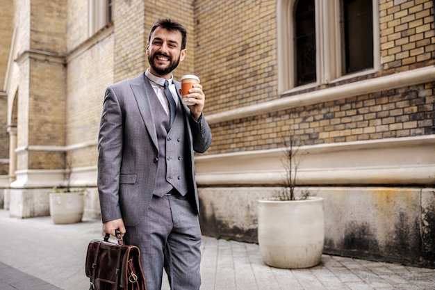 Young attractive bearded businessman holding disposable cup with coffee and going to work.