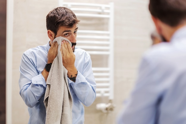 仕事に行く準備をしている若い魅力的なひげを生やしたビジネスマン。彼はバスルームにいて、タオルで顔を拭いています。