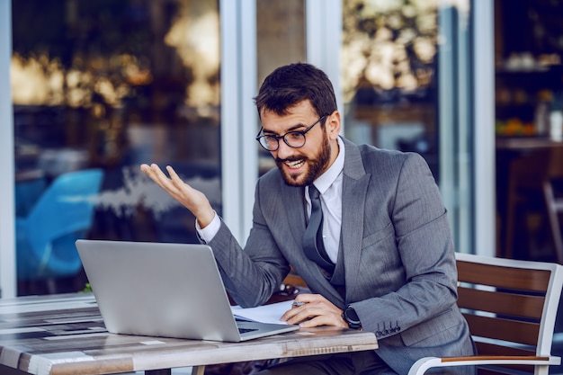 Young attractive bearded smiling caucasian businessman writing tasks in agenda and looking at laptop. Cafe exterior.