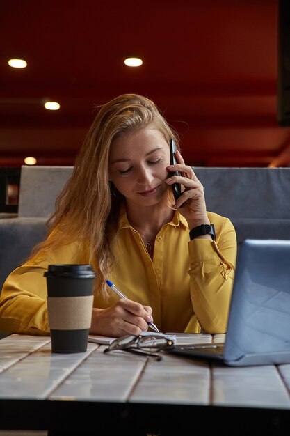 ラップトップに取り組んでいるコーヒーショップに座っている若い魅力的な金髪の白人女性