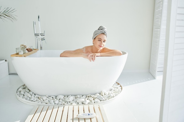 Young attractive blond wet woman in foam with towel on head takes relaxing bath enjoys and smiles Home spa white hotel bathroom interior