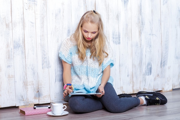 Young attractive blond woman is using tablet in front of white wooden wall