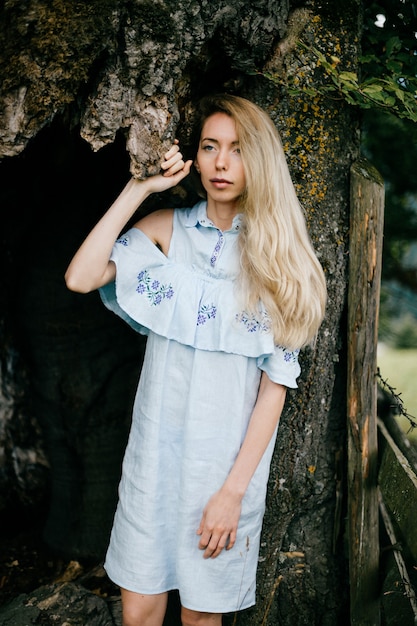 Young attractive blonde girl in blue romantic dress posing with old tree