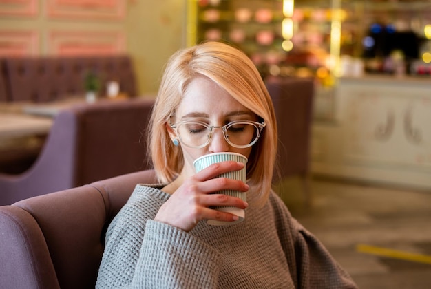 Una giovane ragazza bionda attraente in un caffè beve caffè da una tazza di carta e si rilassa