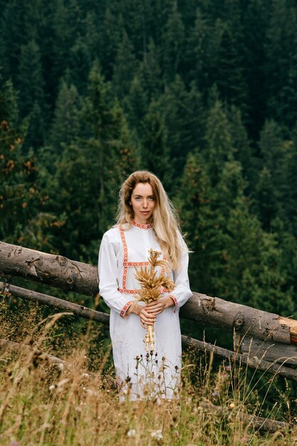 Young attractive blonde girl in white dress with embroidery posing with spikelets bouquet in the meadow over picturesque landscape