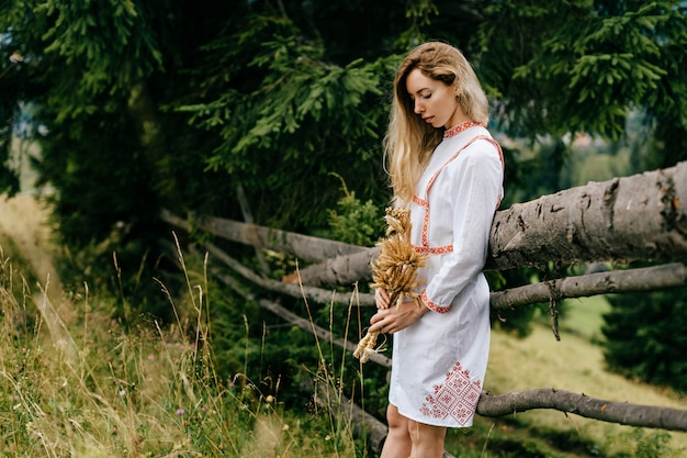 Young attractive blonde girl in white dress with embroidery posing with spikelets bouquet near wooden fence