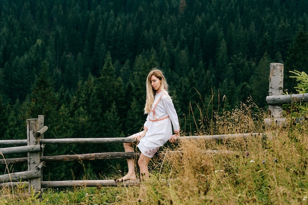 Young attractive blonde girl in white dress with embroidery sitting on wooden fence over picturesque forest landscape