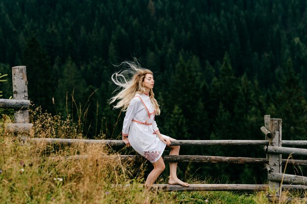 Foto giovane ragazza bionda attraente in vestito bianco con ricamo che si siede sulla staccionata in legno sul pittoresco paesaggio forestale